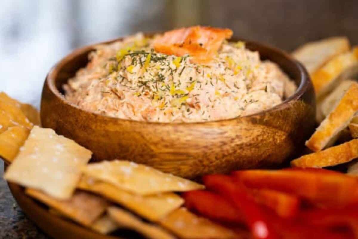 A wooden bowl filled with dip, surrounded by crackers and sliced bell peppers.