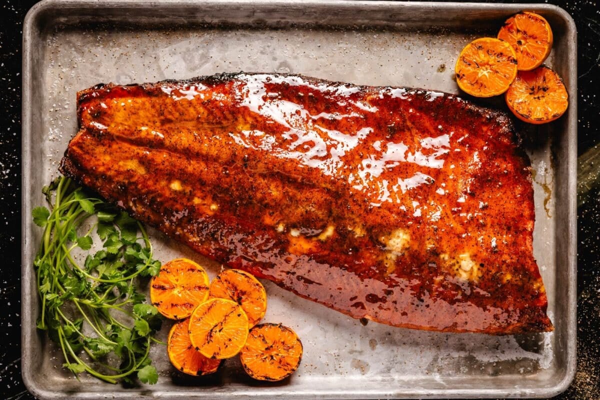 A glazed salmon fillet on a baking sheet with orange slices and herb garnish.