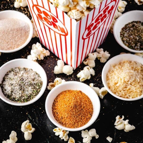 Five separate seasoning blends in bowls on a counter with a carton of popcorn.