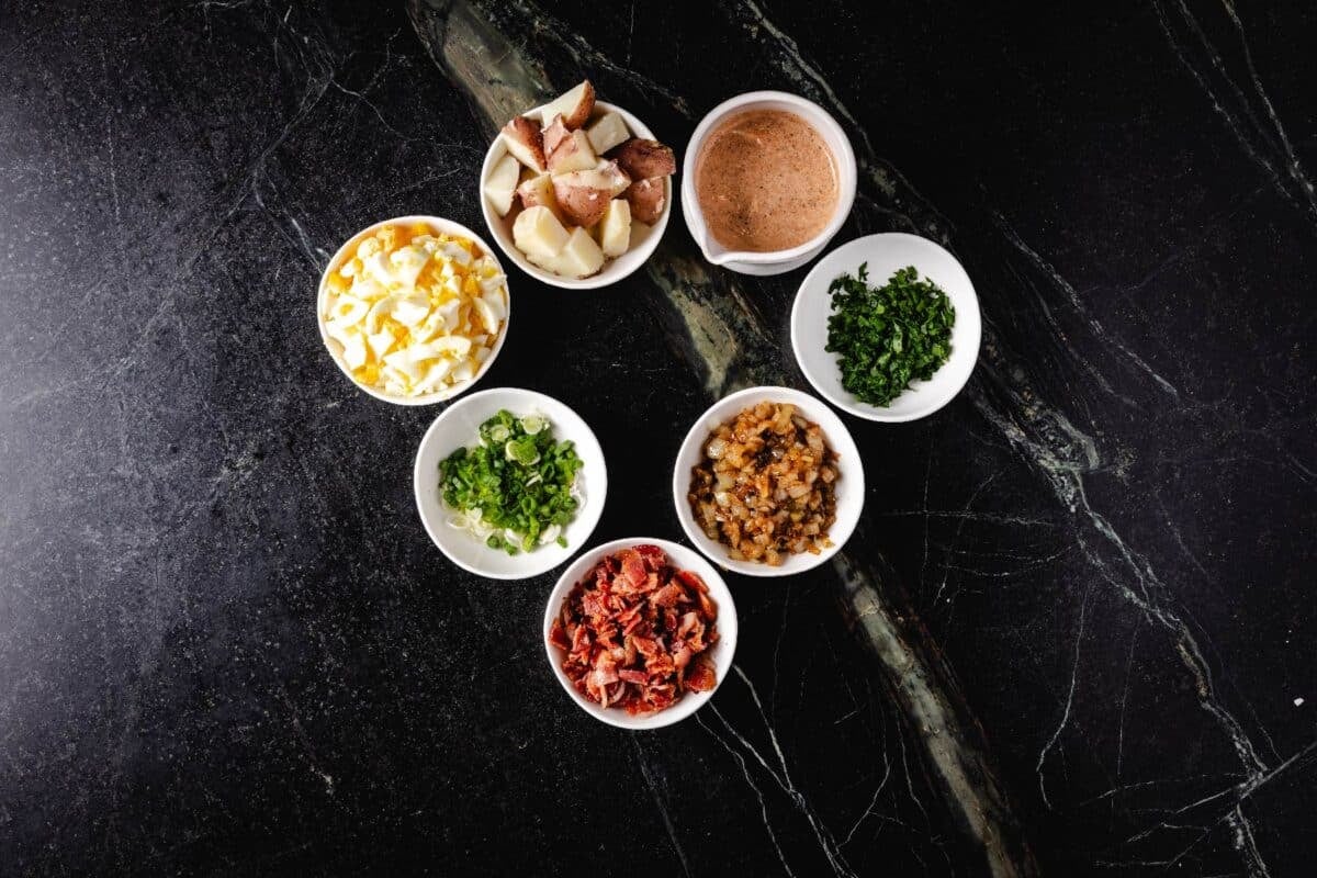 Chopped red potatoes, eggs, bacon, herbs, and other ingredients in white bowls on a marble surface.