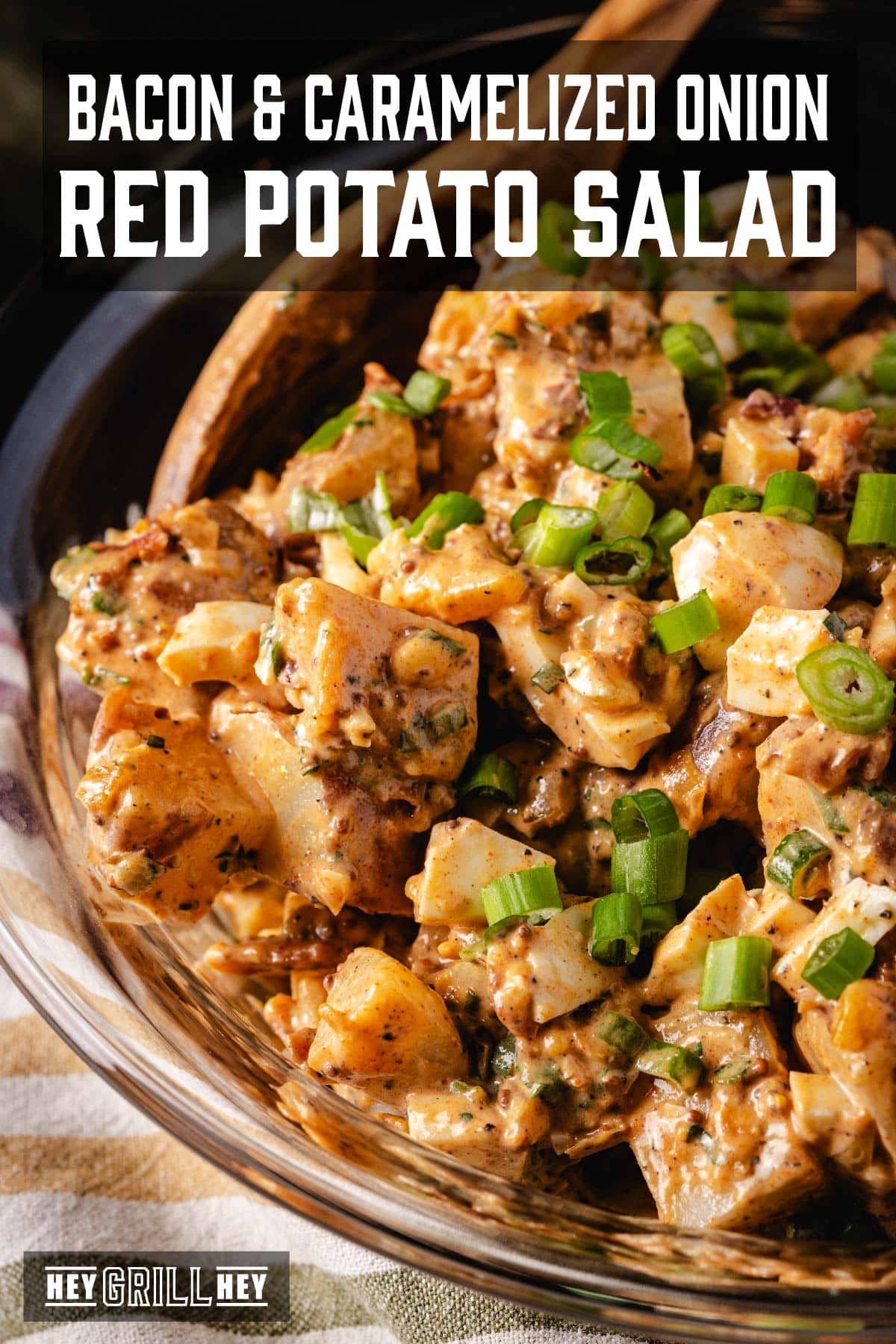 A glass bowl full of potato salad, with a wooden spoon resting in it. The text overlay reads "Bacon and Caramelized Onion Red Potato Salad" at the top, and "Hey Grill Hey" at the bottom.