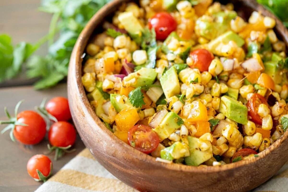 A wooden bowl filled with a recipe for corn salad, next to some cherry tomatoes.