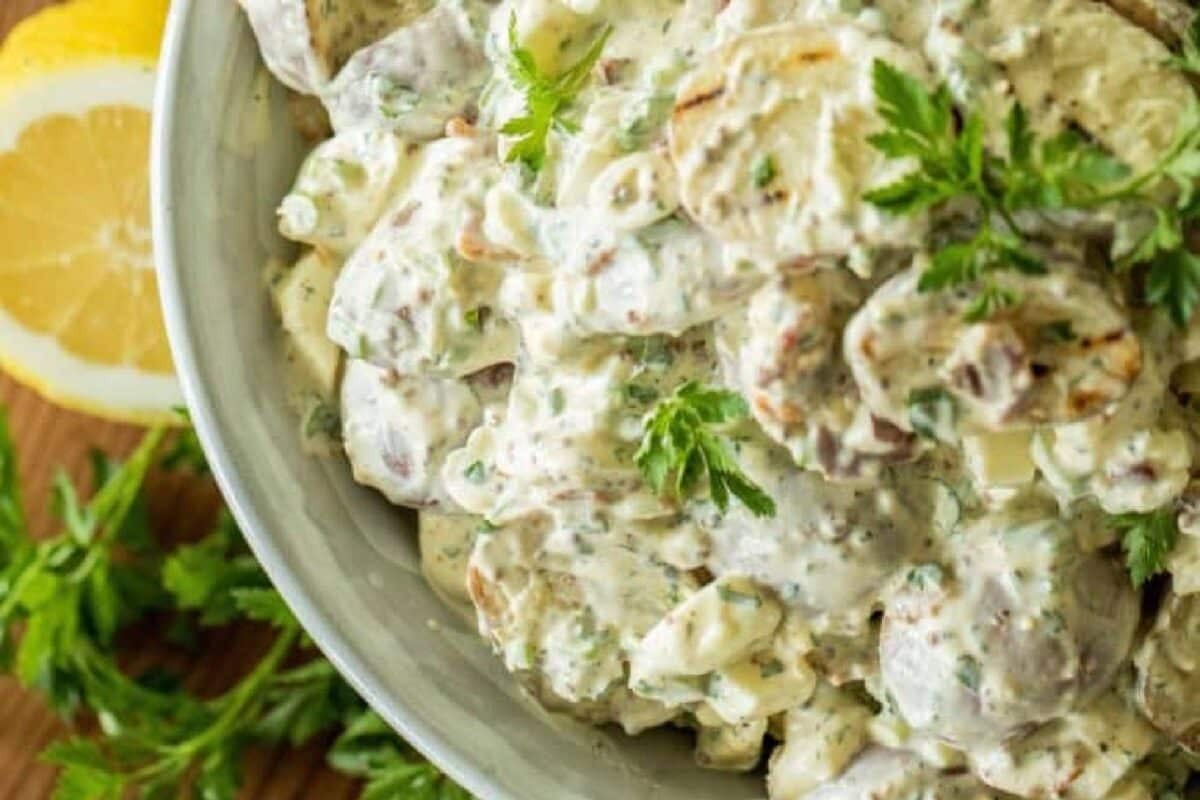 A potato salad recipe in a white bowl next to a lemon wedge and herb garnish.