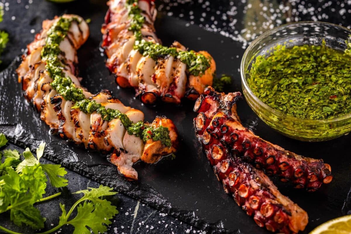 Grilled and sliced octopus on a serving platter next to a bowl of sauce for seafood.