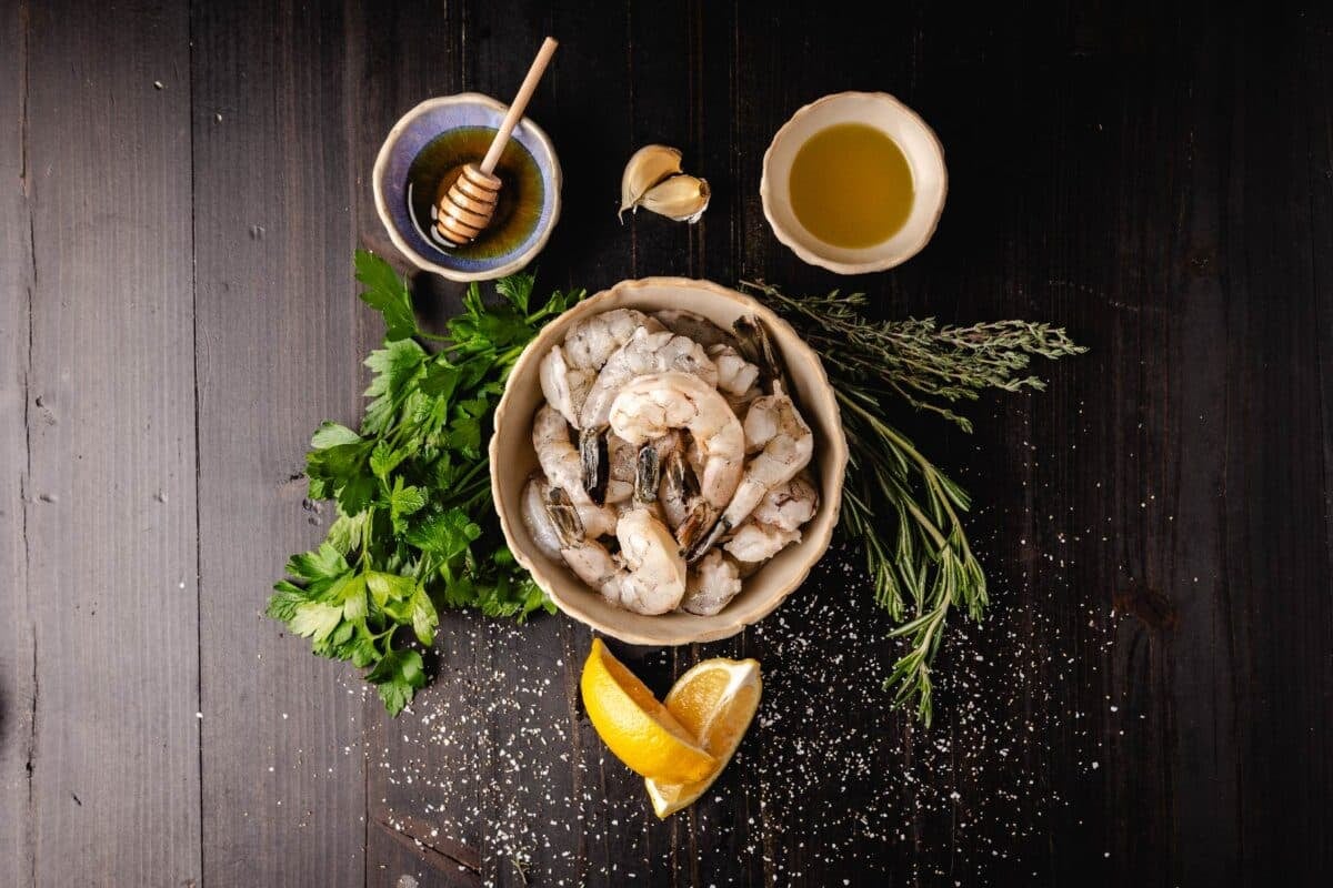 A bowl of raw shrimp on a black countertop surrounded by the recipe ingredients for a seafood marinade.