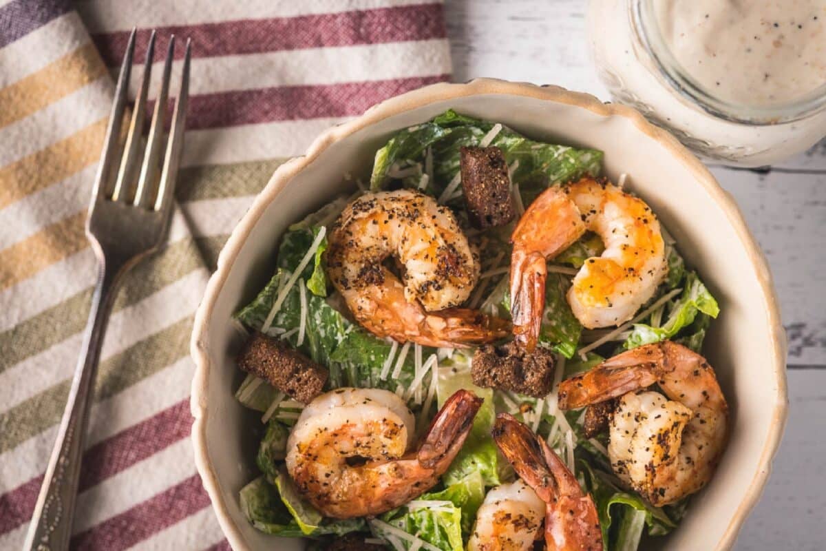 A salad topped with grilled shrimp, sitting on a tablecloth next to a fork.