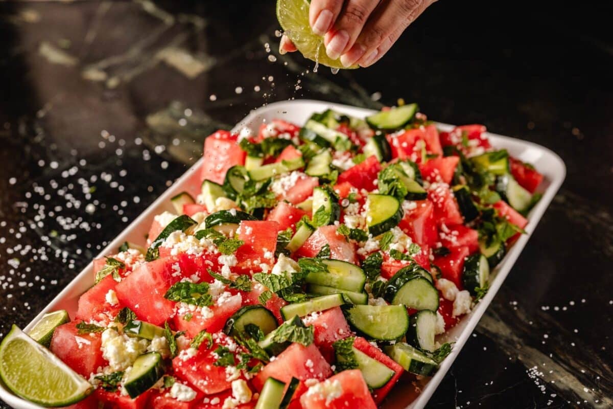 Sliced watermelon and cucumber on a large platter, with a hand squeezing fresh juice from a lime onto them.