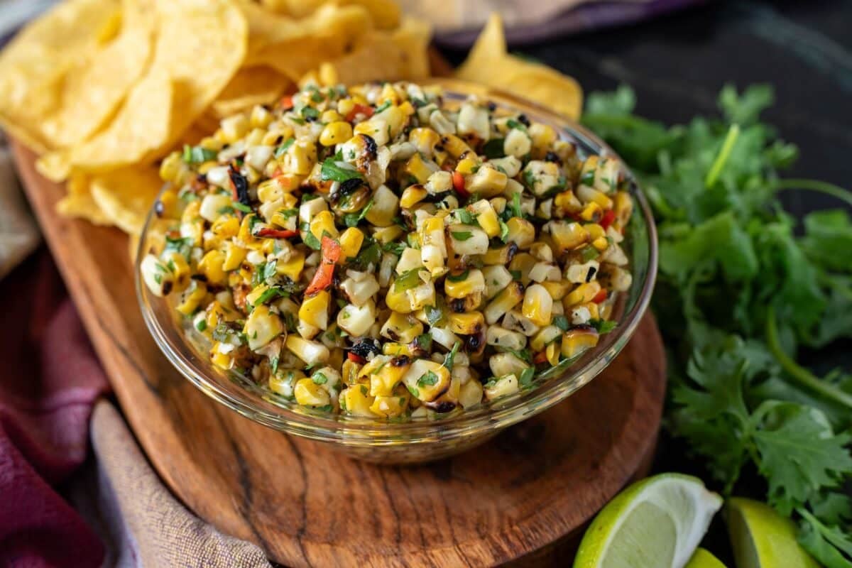 Corn salsa in a glass bowl on a wooden platter with tortilla chips and lime wedges.