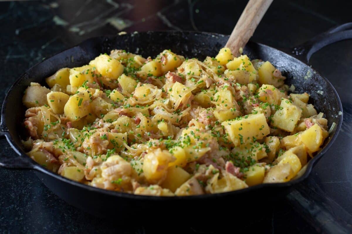 A wooden spoon stirring potatoes and bacon in a cast iron skillet.