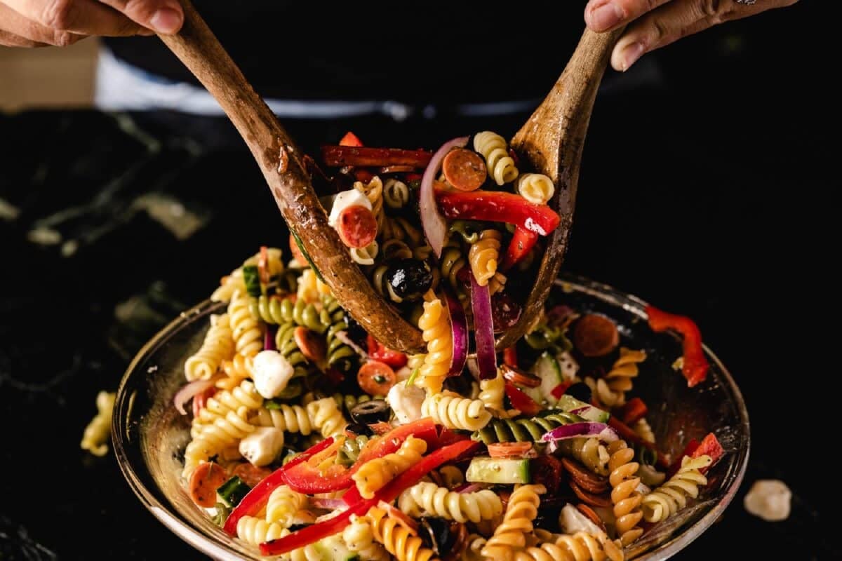 Wooden serving tools tossing a pasta salad in a glass bowl.