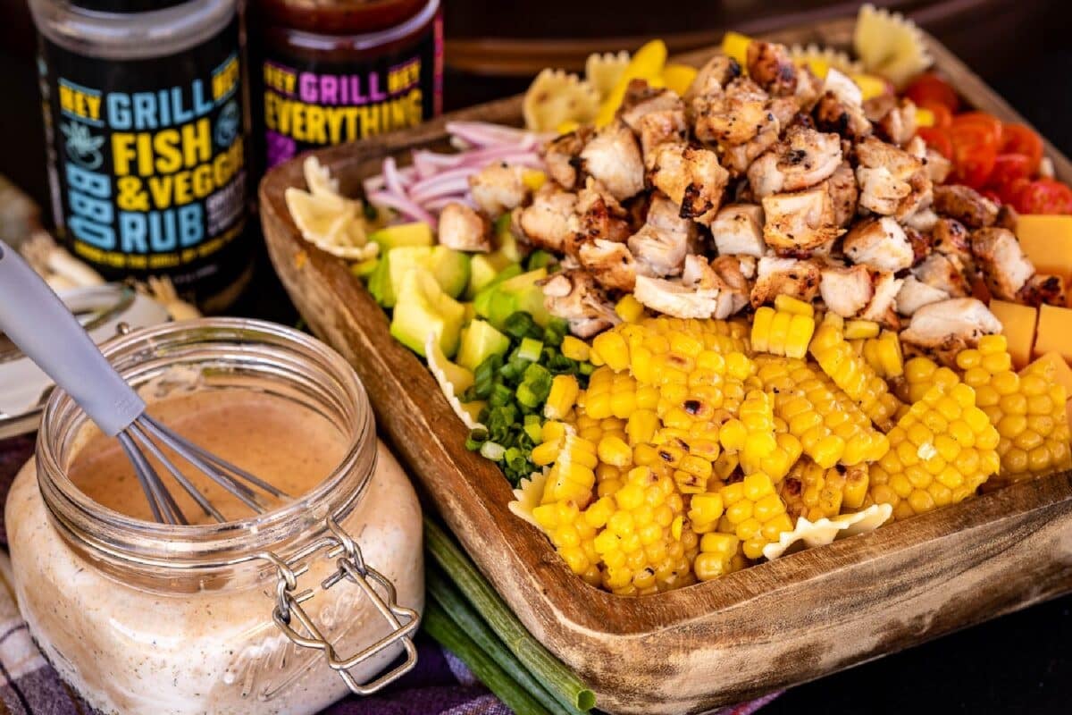 Grilled chicken, corn, and other ingredients on a wooden cutting board next to a jar of sauce.