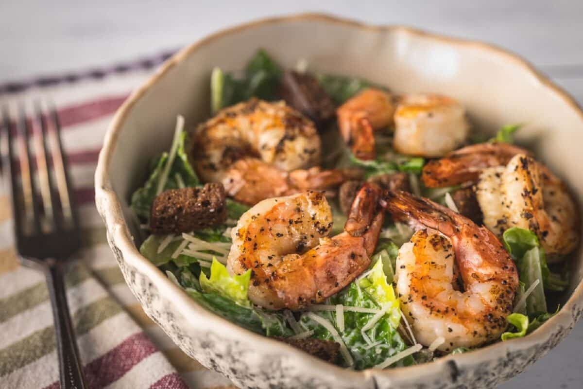 A green salad topped with shrimp in a bowl, on a tablecloth next to a fork.