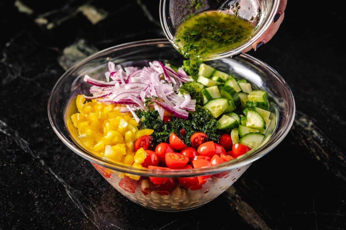 A glass bowl of dressing being poured into a larger glass bowl full of chopped veggies.