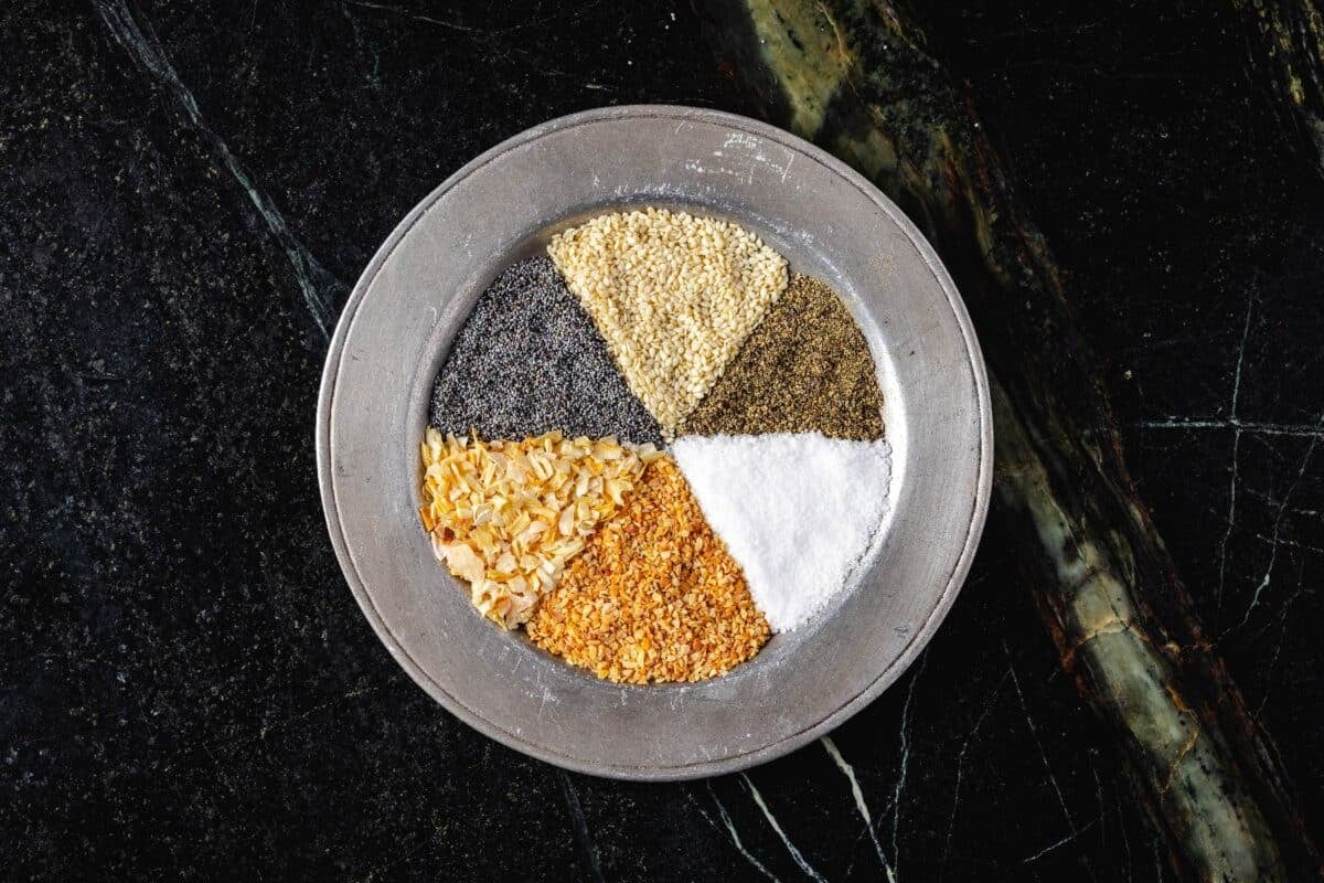 A metal plate with equal portions of dried onion, dried garlic, sesame seeds, poppy seeds, salt, and pepper formed into triangles.