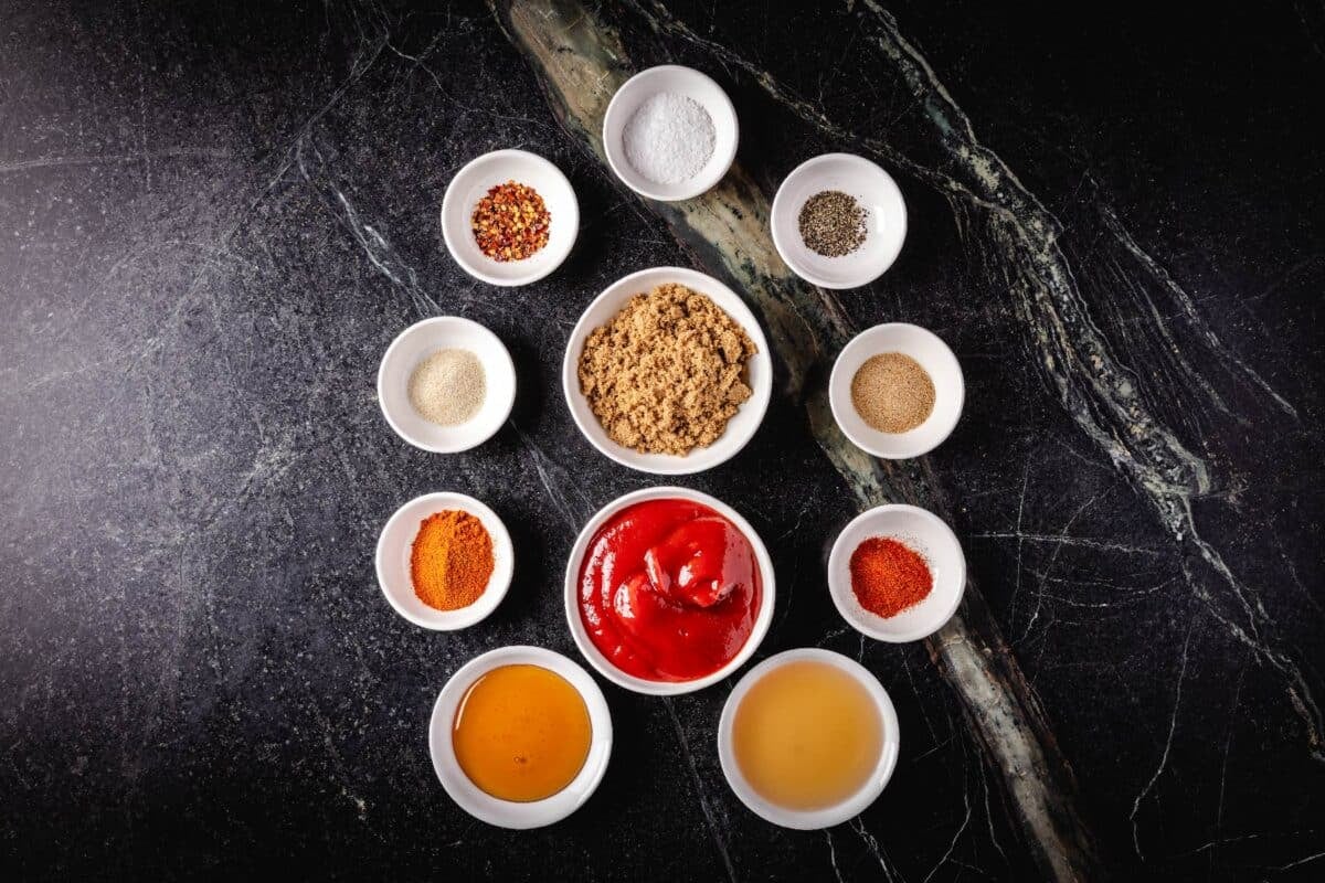 An overhead shot of glass bowls filled with ketchup, brown sugar, and various spices on a marble surface.