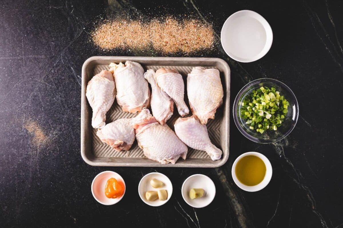 Raw chicken legs on a baking sheet next to bowls of marinade ingredients, and a pile of jerk seasoning.