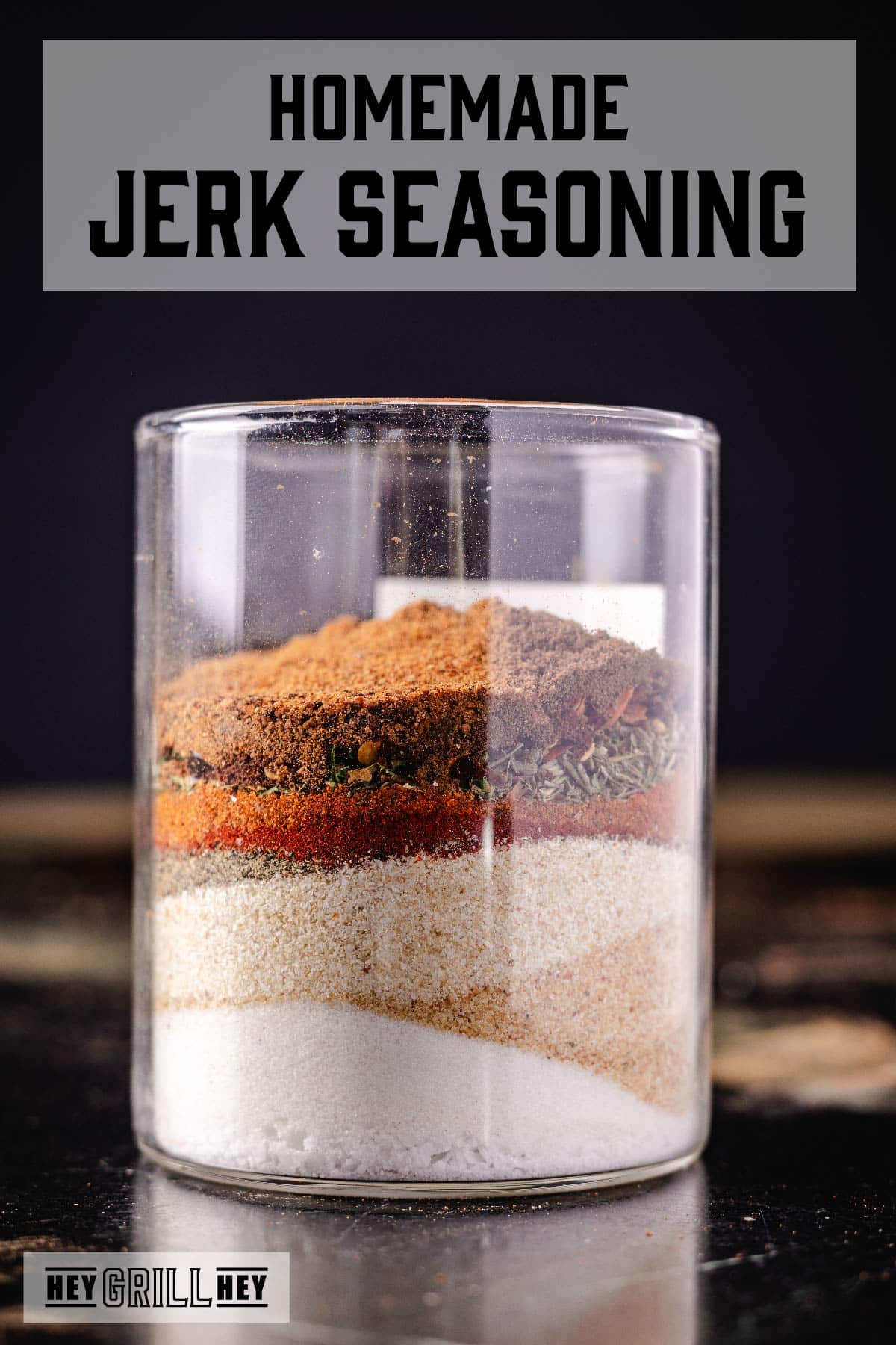 A glass jar with various seasonings layered inside it. The text overlay reads "Homemade Jerk Seasoning" at the top, and "Hey Grill Hey" at the bottom.