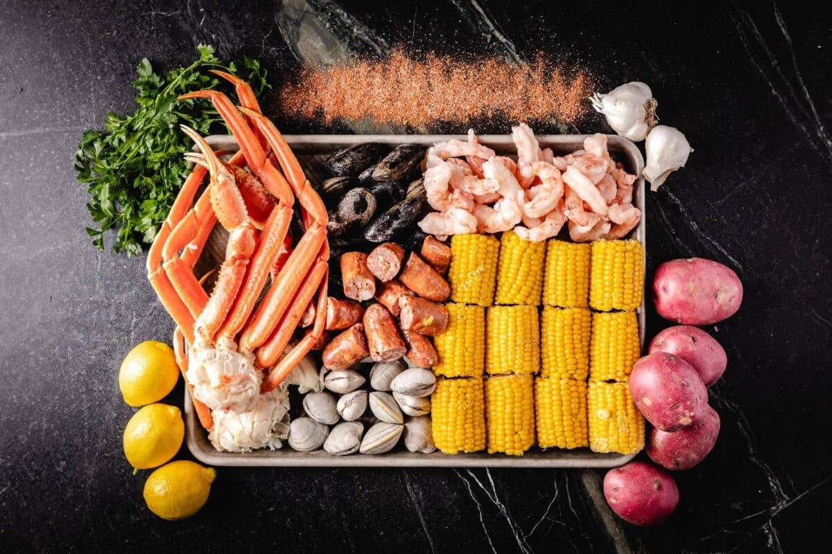 A baking sheet with corn cob sections and various types of seafood on it. There are lemons, potatoes, parsley, and a small pile of Old Bay seasoning around it.