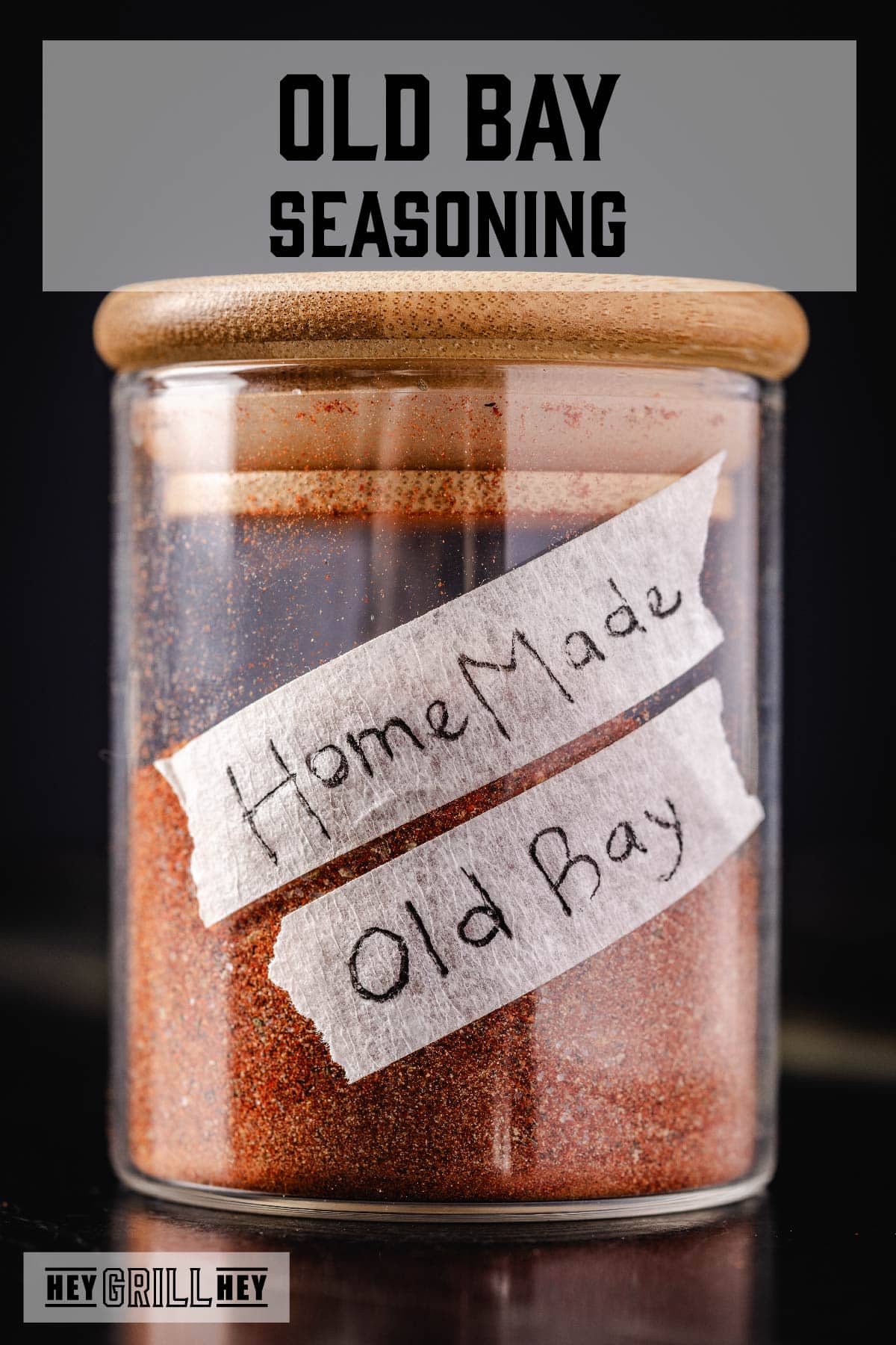 A glass gar of seasoning with a wooden lid. There are two pieces of tape on the jar with "Homemade Old Bay" written on them. The text overlay reads "Old Bay Seasoning" at the top, and "Hey Grill Hey" at the bottom.