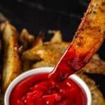 A potato wedge being dipped into a while cup of ketchup, with more wedges in the background.