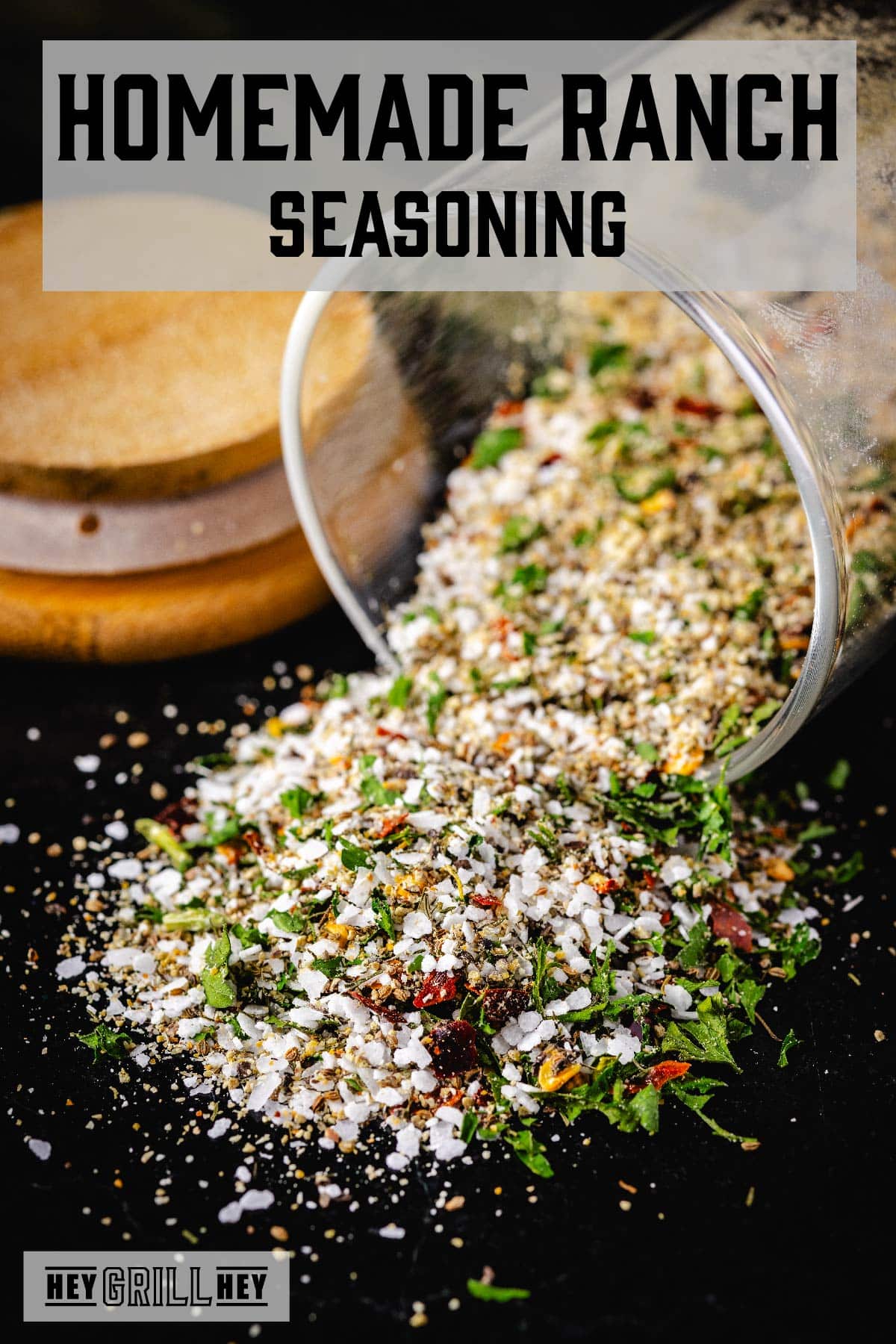 A glass jar tipped with seasoning spilling out, in front of a wooden lid. The text overlay reads "Homemade Ranch Seasoning" at the top, and "Hey Grill Hey" at the bottom.