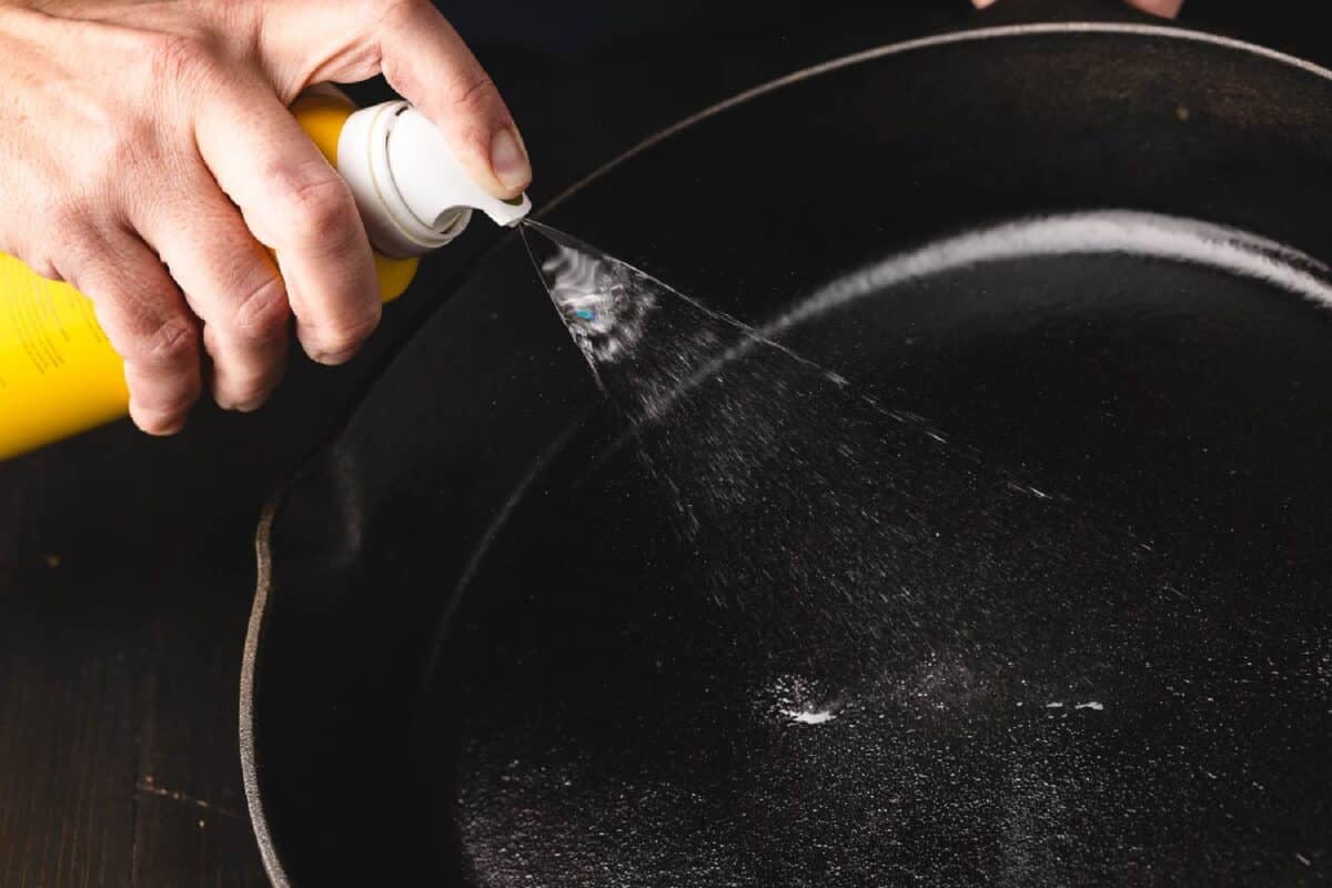 A hand spraying seasoning oil onto a cast iron pan.