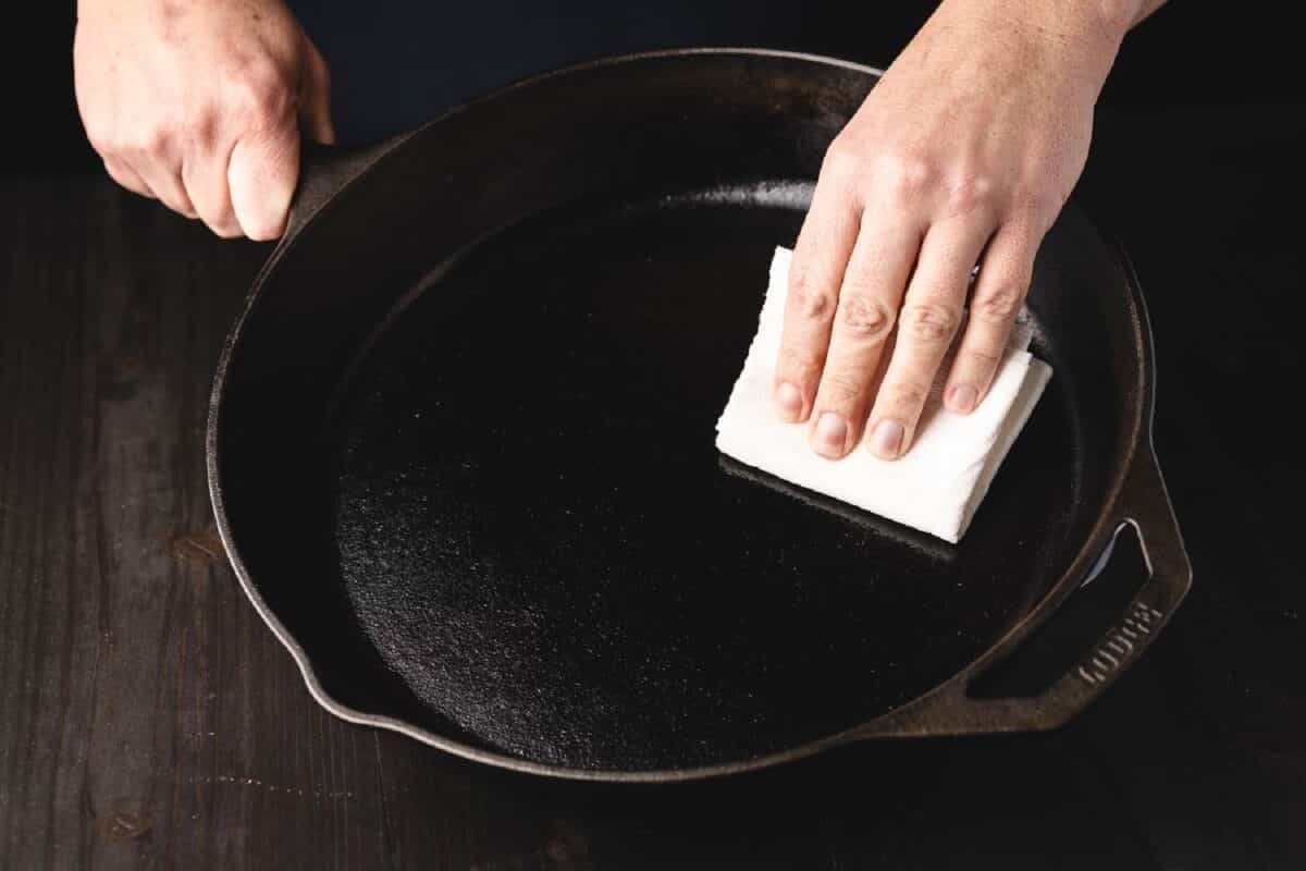 A hand using a paper towel to rub seasoning oil into a cast iron skillet.