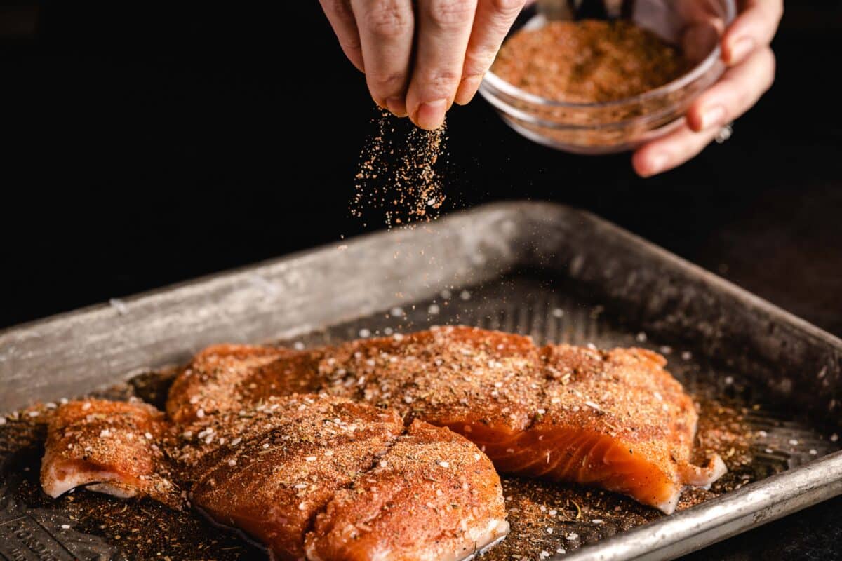 Salmon fillets on a baking sheet with a hand sprinkling a spice blend onto them from a small bowl.