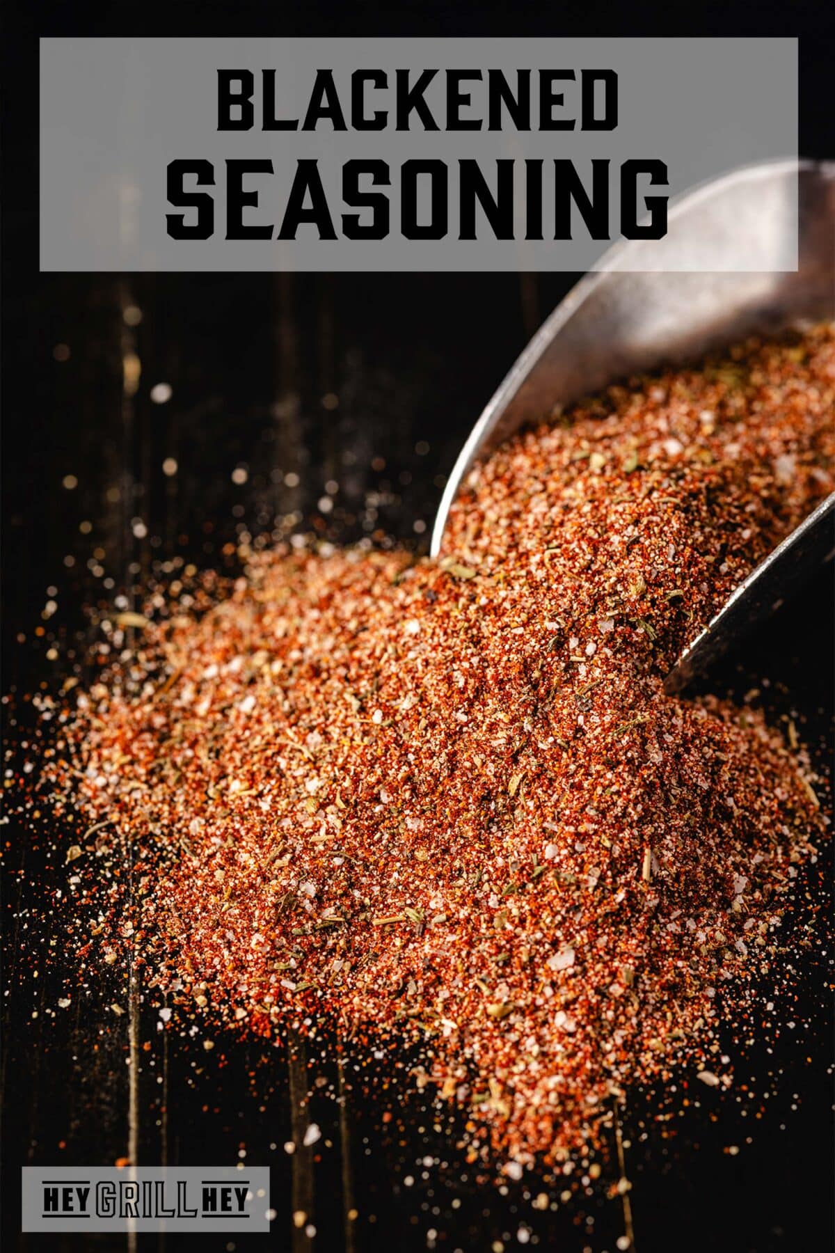 A metal scoop resting in a pile of seasoning on a black countertop. The text overlay reads "Blackened Seasoning" at the top, and "Hey Grill Hey" at the bottom.