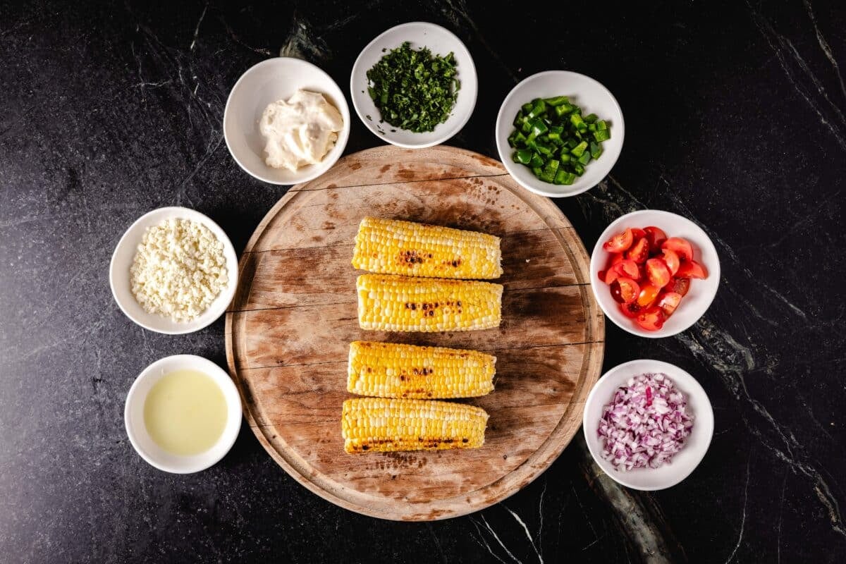 Grilled corn on a round, wooden cutting board, surrounded by 7 white bowls of various seasonings and sliced veggies.