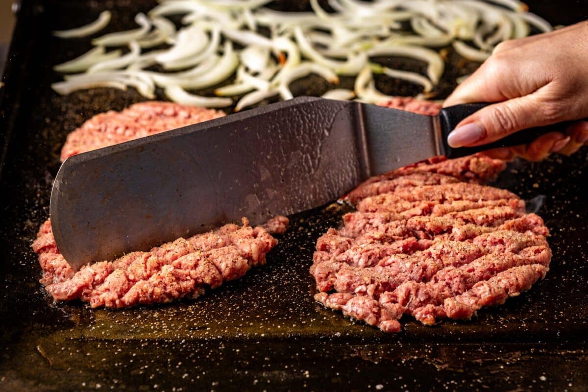 A spatula cutting into beef patties on a flat-top grill with sliced onions in the background.