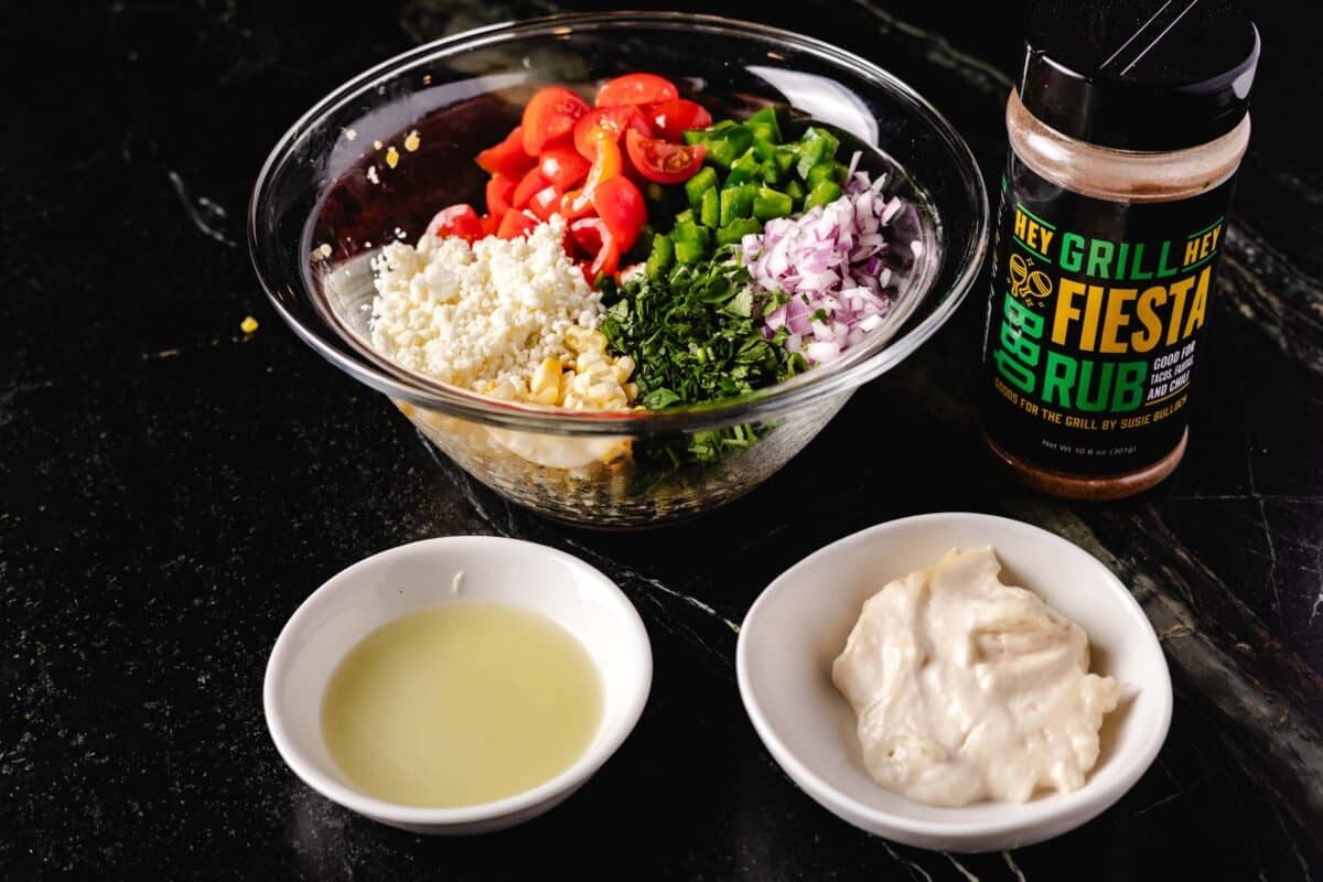 A large glass bowl filled with various chopped veggies next to a bottle of BBQ seasoning, a small white bowl with oil, and another small bowl with mayo.