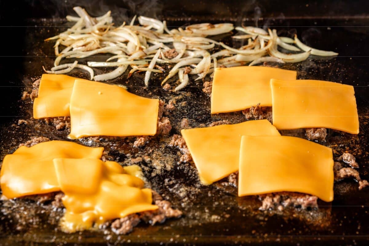 Ground beef topped with cheese slices, and sliced onions on a flat-top grill.