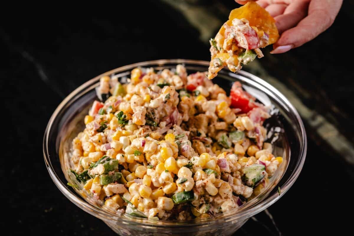 A large glass bowl full of corn salsa, and a hand holding a chip with a scoop of the dip above the bowl.