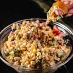 A large glass bowl full of corn salsa, and a hand holding a chip with a scoop of the salsa above the bowl.