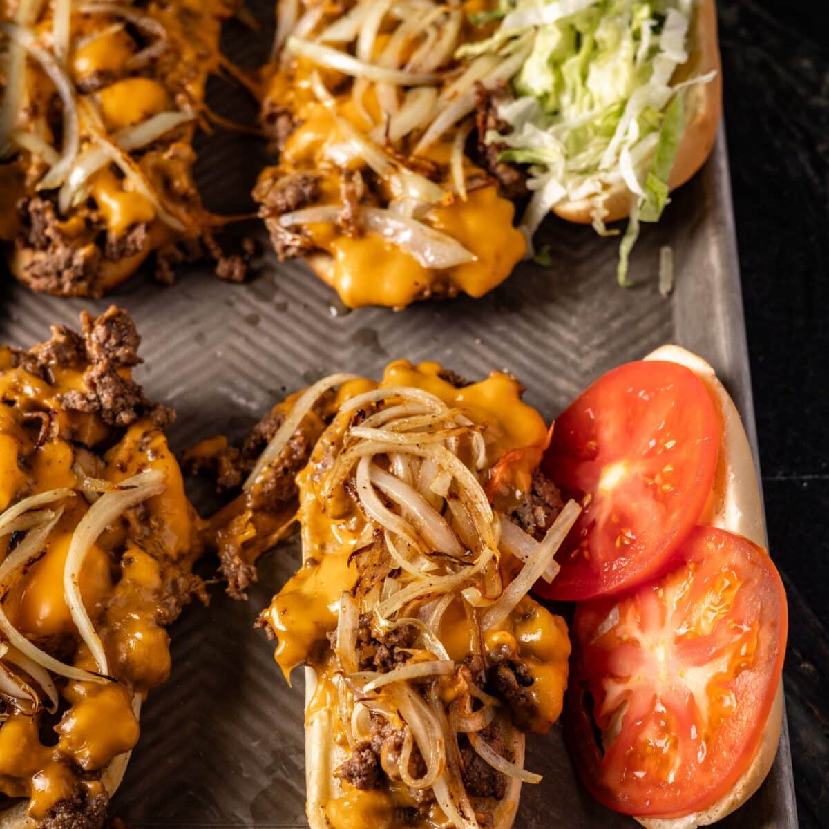 Several hoagies on a baking sheet with beef, cheese, tomatoes, and shredded lettuce.