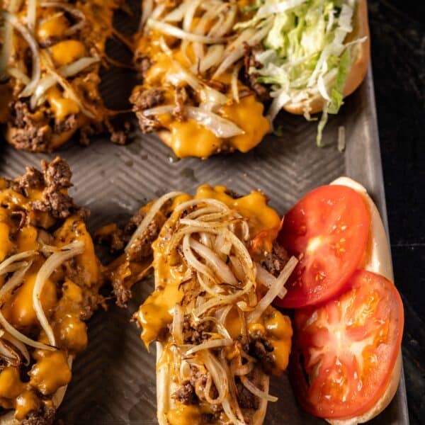 Several hoagies on a baking sheet with beef, cheese, tomatoes, and shredded lettuce.