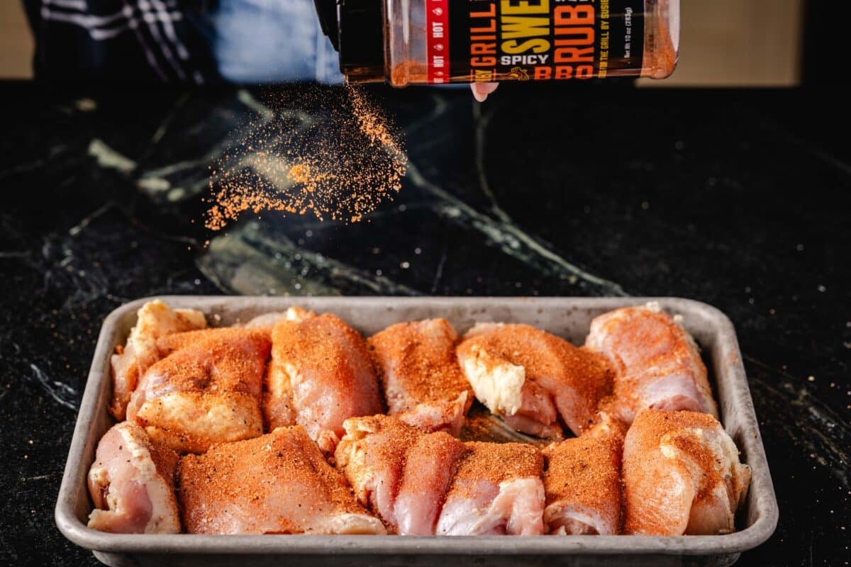 Chicken thighs on a baking sheet with a hand above sprinkling them with BBQ rub.