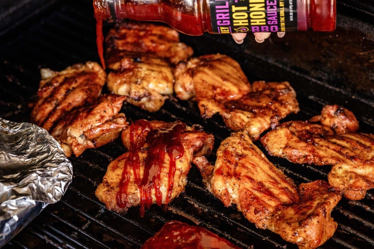 Poultry sitting on smoker grates being drizzled with BBQ sauce.