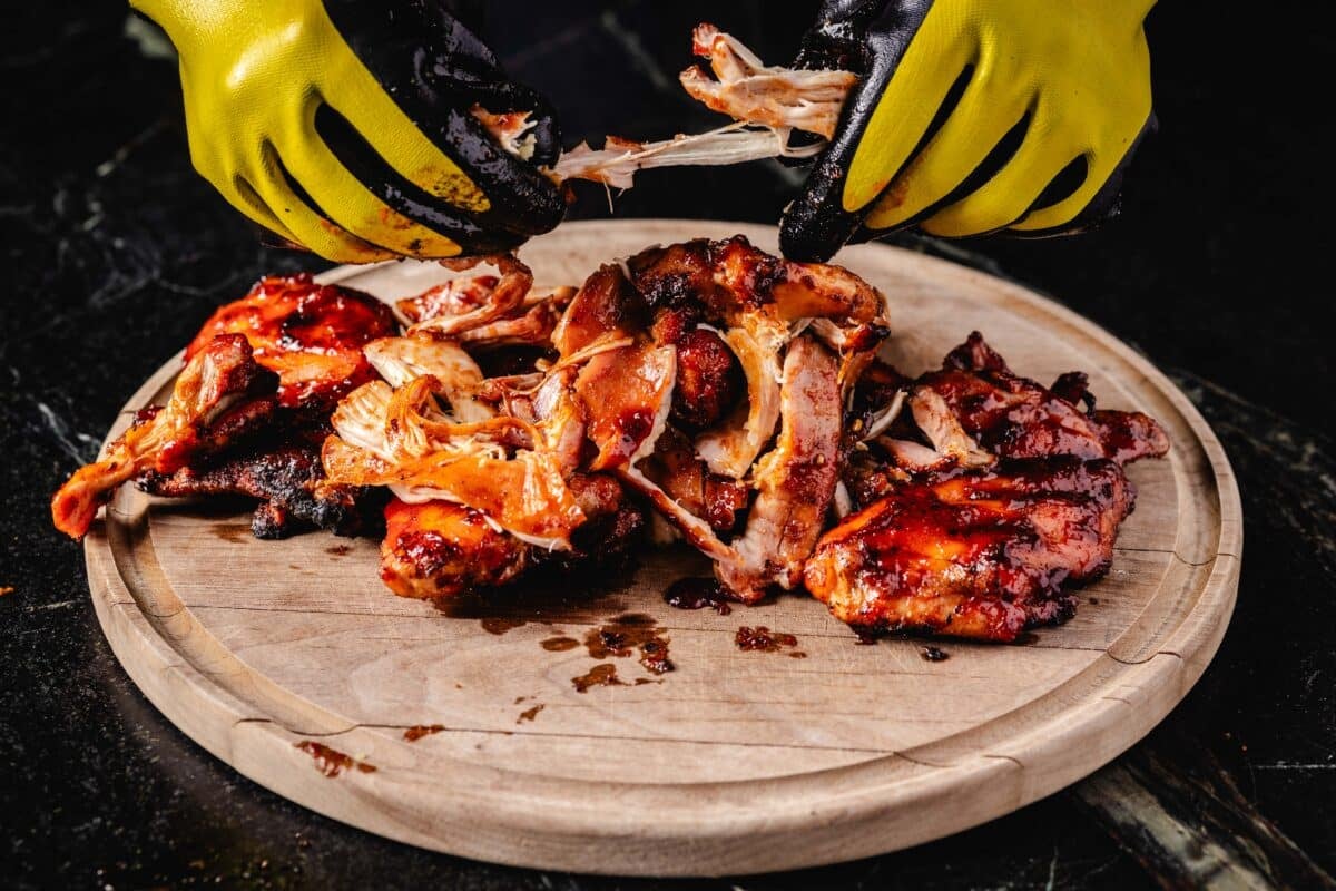 Gloved hands shredding smoked chicken on a wooden cutting board.