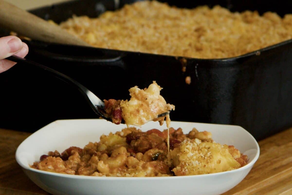 A fork holding a scoop of homemade chili mac above a bowl of it.