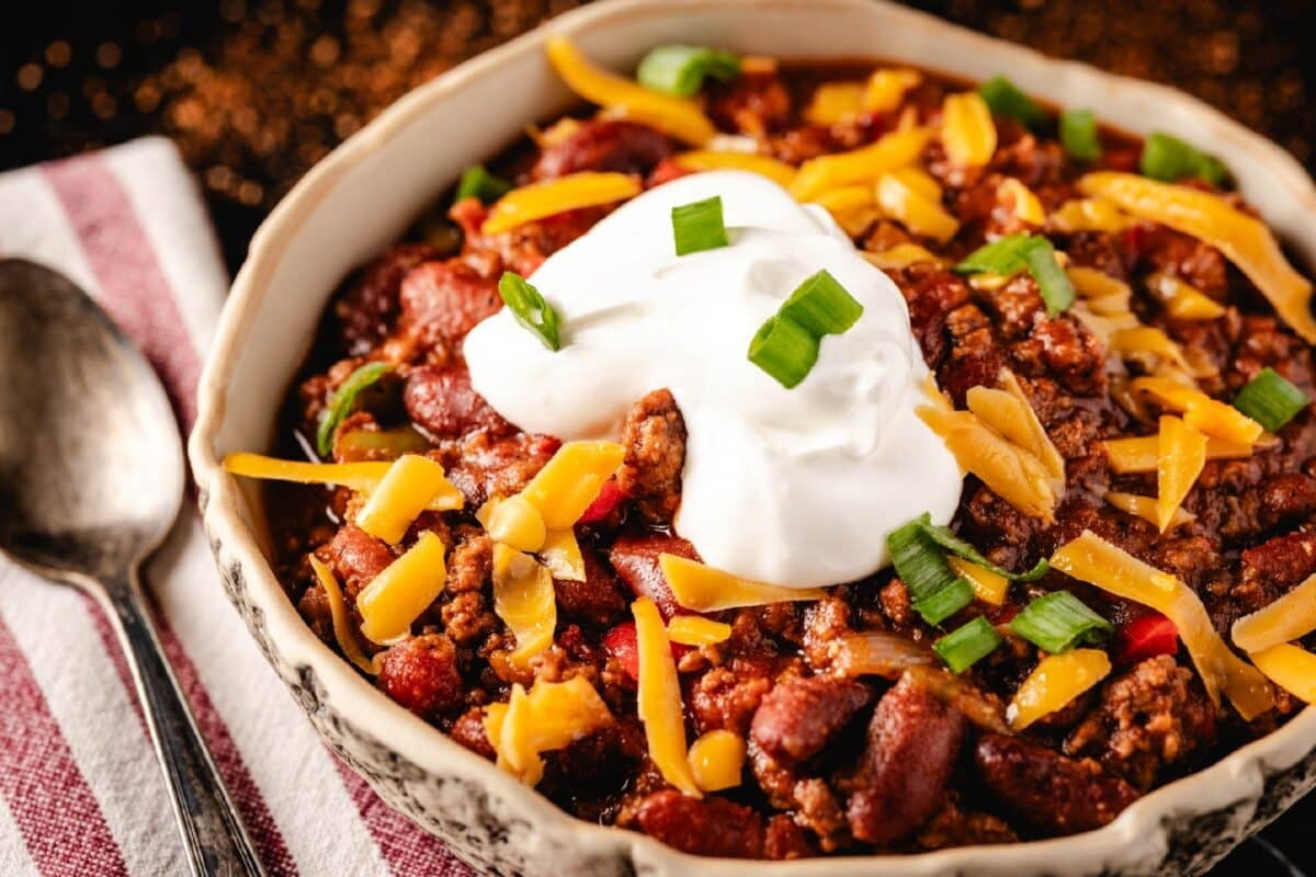 Homemade ground beef chili in a white bowl next to a spoon and tablecloth.