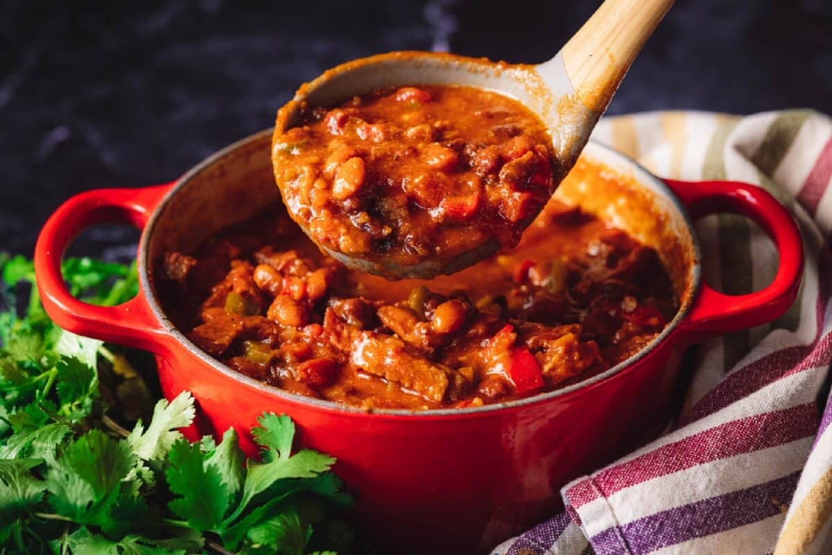 A tri tip chili recipe in a pot with a wooden spoon scooping some out.