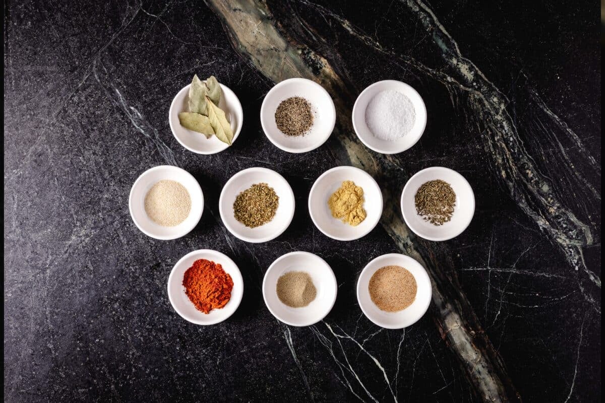 Ten white bowls in 3 rows (3, 4, 3) fulled with various spices on a marble surface.