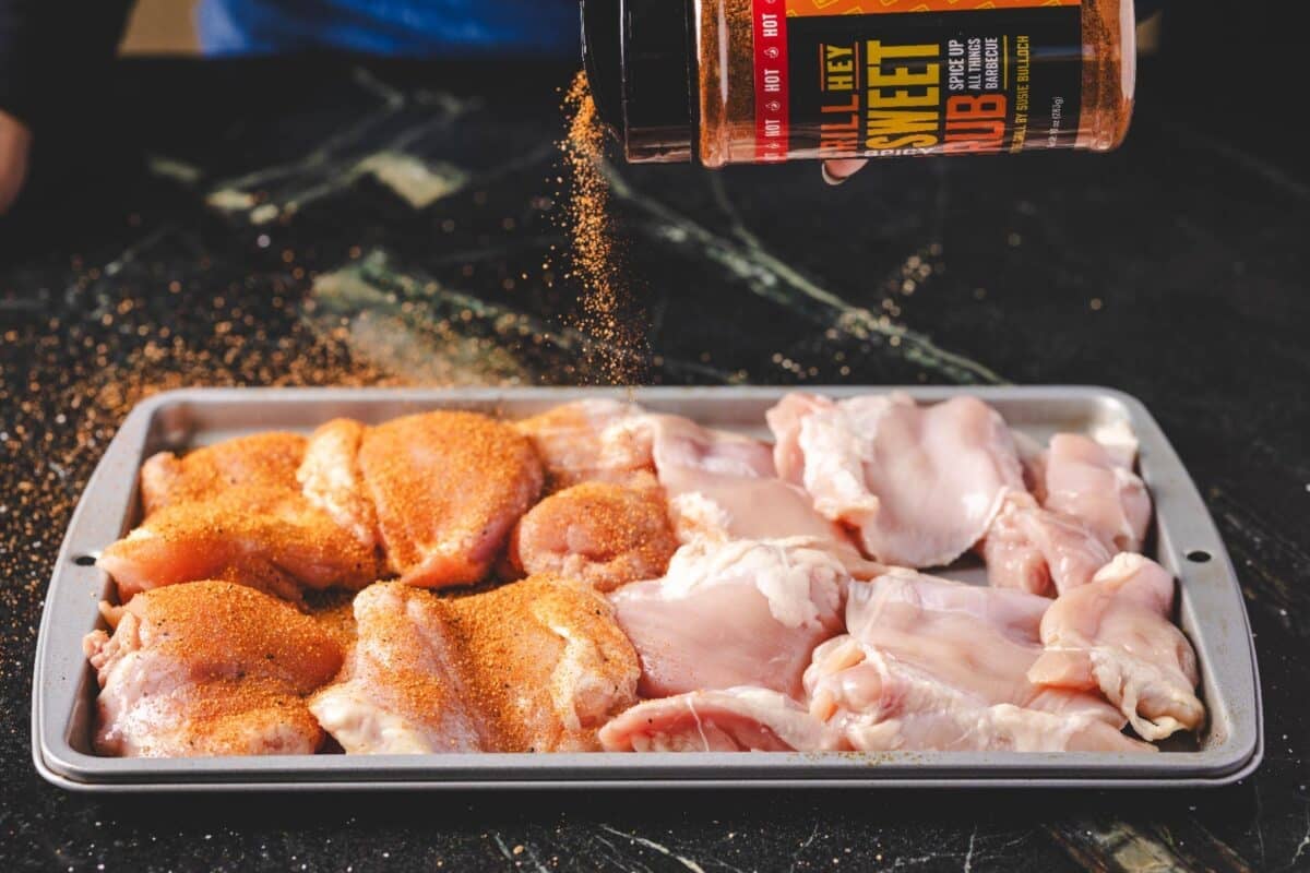 Poultry breasts on a baking sheet being sprinkled with Spicy Sweet Rub from a bottle.