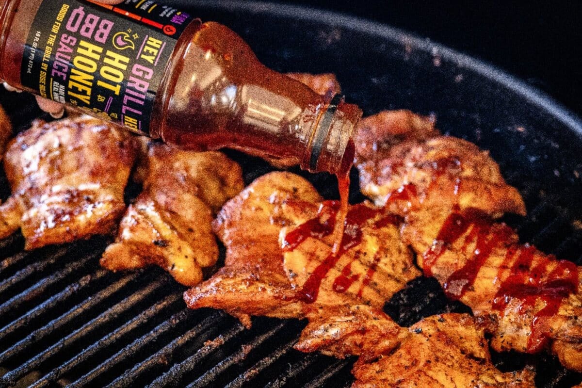 A bottle of Hot Honey BBQ Sauce pouring onto chicken breasts sitting on grill grates.