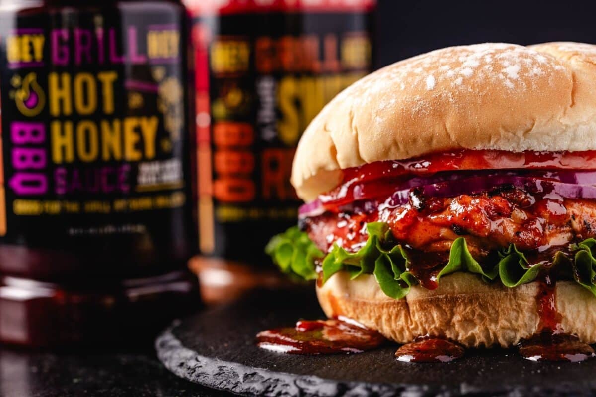 A grilled chicken sandwich with lettuce, tomato, and onion, in front of a bottle of BBQ sauce and a bottle of BBQ rub.