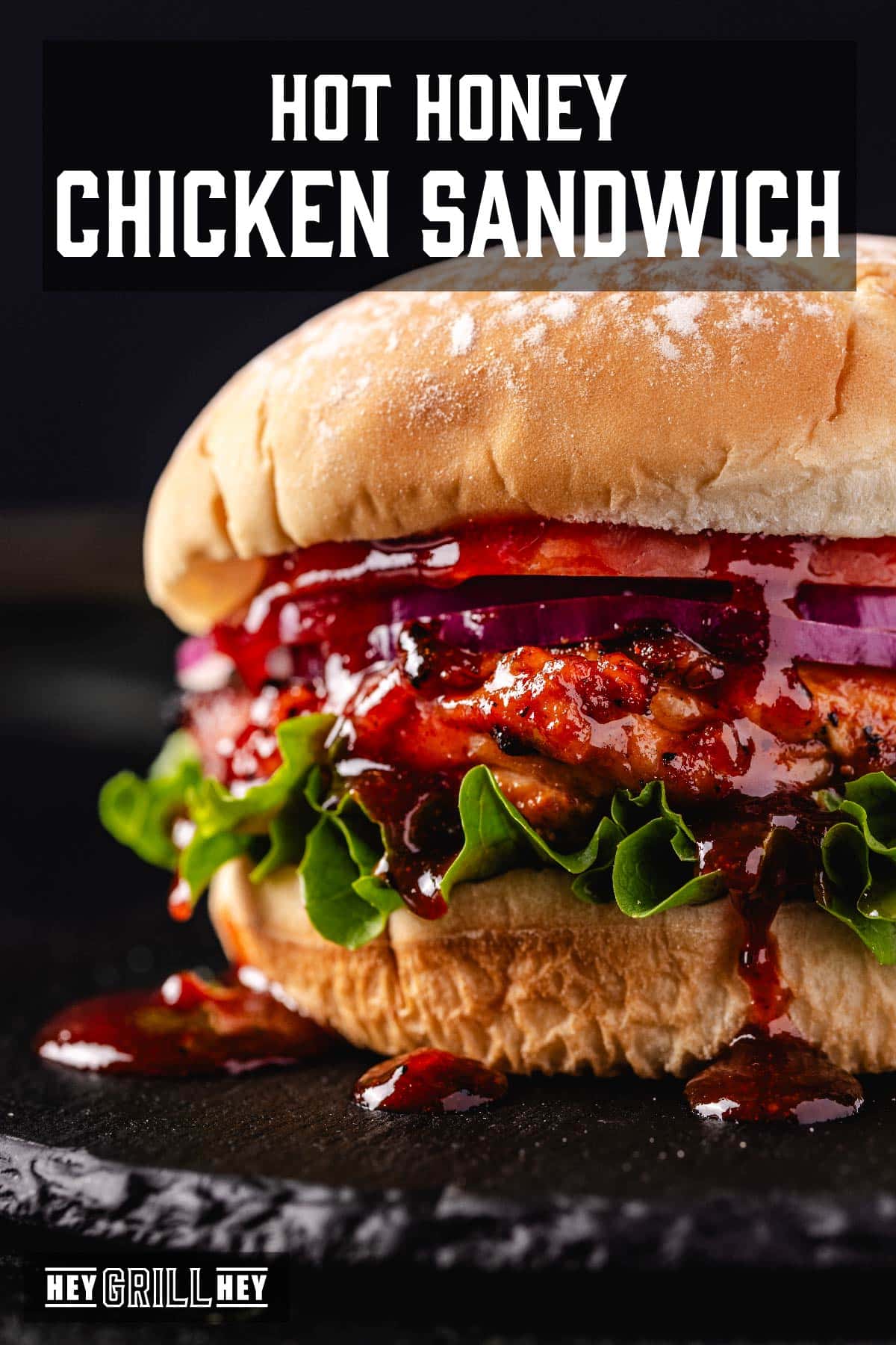 A grilled chicken sandwich with lettuce, tomato, and onion, dripping with BBQ sauce on a black stone platter. The text overlay reads "Hot Honey Chicken Sandwiches" at the top, and "Hey Grill Hey" at the bottom.