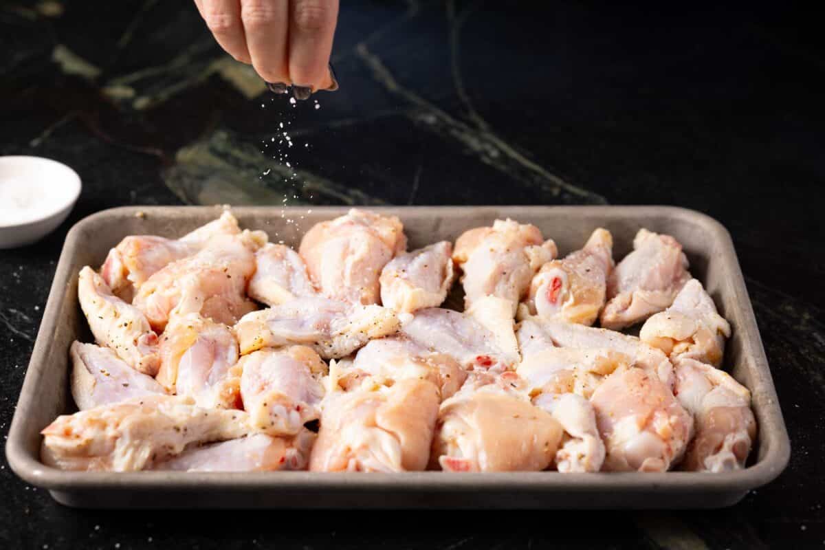 Raw chicken flats and drumettes on a baking sheet as a hand above sprinkles salt on them.