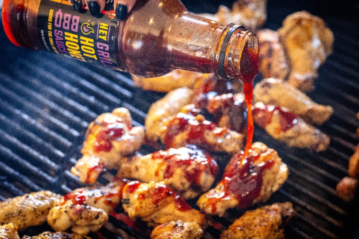 Chicken wings sitting on grill grates as a hand pours Hot Honey BBQ Sauce onto them.